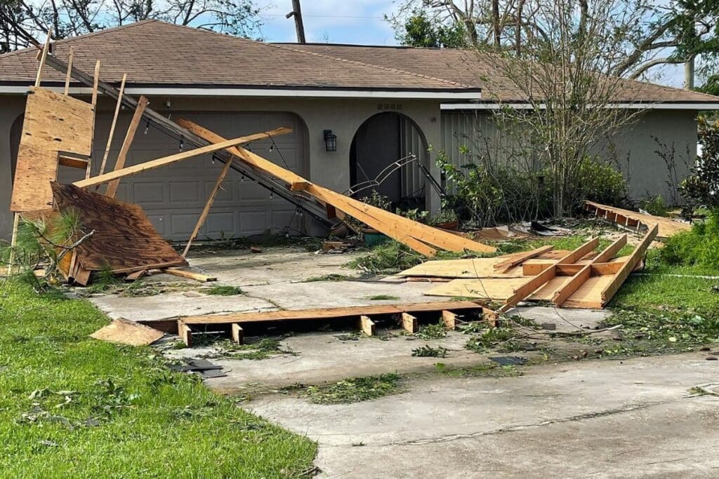 House with visible hurricane damage, including debris and collapsed wooden structure in the driveway.